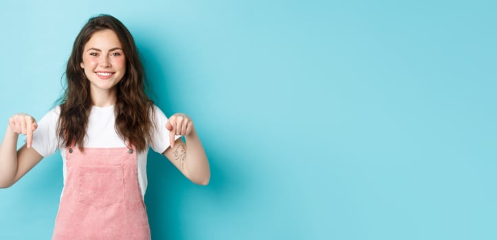 Place your logo here. Cheerful brunette woman pointing fingers down and smiling at camera, recommending product, showing sale promo offer, standing against blue background photo