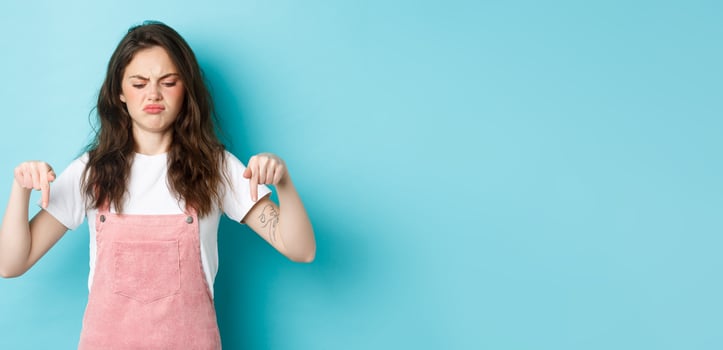 Sounds bad. Displeased grumpy brunette girl frowning and sulking moody, pointing fingers down and look disappointed, standing against blue background photo