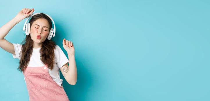 Beautiful young woman dancing and listening music in headphones, enjoying sound, standing over blue background photo