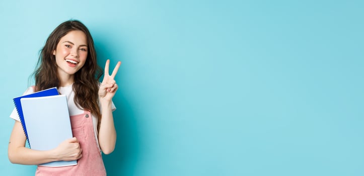 Beautiful female student showing v-sign and smiling happy, holding notebooks with study material, attending courses, standing over blue background photo