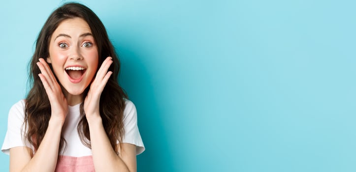 Close up of excited young woman holding hands near face, gasping and looking amazed at camera, stare surprised at super cool news, standing over blue background photo
