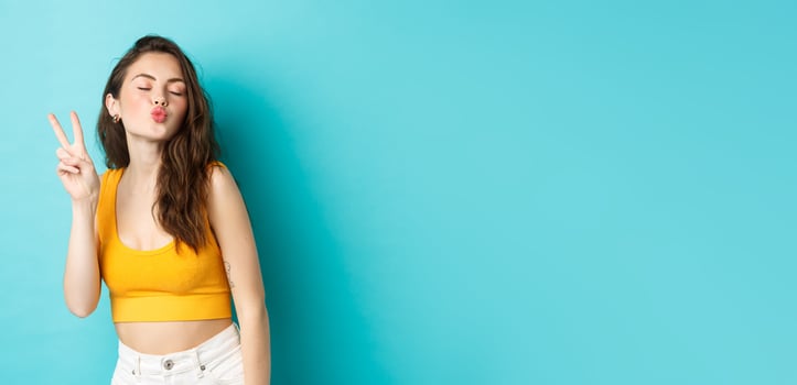 Glamour girl in summer clothes close her eyes and making kissing face with v-sign, enjoying vacation, having fun, standing relaxed against blue background photo
