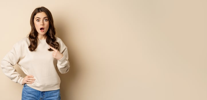 Surprised girl pointing at herself and look with disbelief, standing over beige background photo