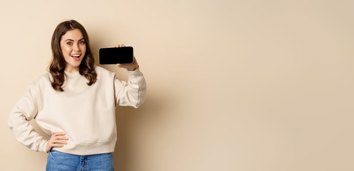 Smiling beautiful female model demonstrating horizontal smartphone screen, mobile application, standing over beige background photo
