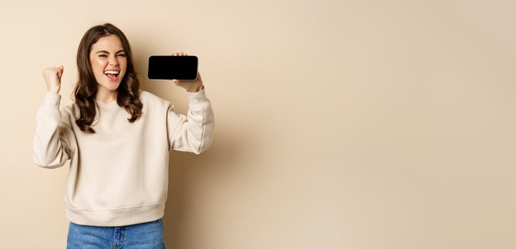 Happy girl showing mobile phone horizontal screen and celebrating, chanting with joyful face, standing over beige background photo