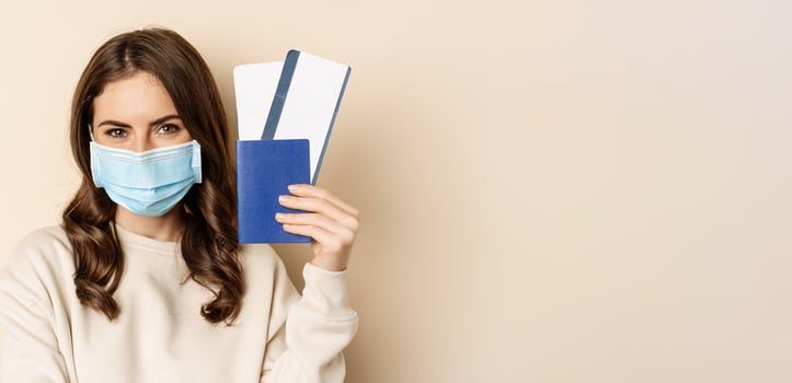 Travel and covid-19 pandemic. Girl in medical face mask travelling, showing passport with two tickets on plane, standing against beige background photo
