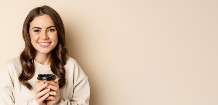 Beautiful authentic woman smiling, holding warm cup of coffee and looking happy, standing over beige background photo