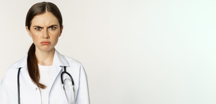 Angry and upset young woman doctor, female healthcare worker sulking, frowning disappointed, dislike smth, standing over white background photo