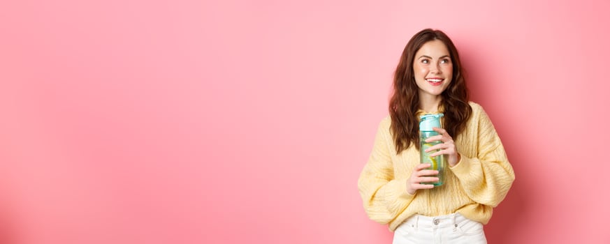 Smiling healthy and sporty brunette girl, looking thoughtful aside at logo, holding water bottle with lemon drink, standing against pink background photo