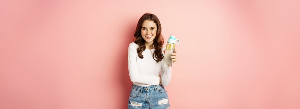 Healthy lifestyle. Happy smiling woman holding water bottle with lemon, vitamin c drink, looking joyful at camera, posing against pink background photo