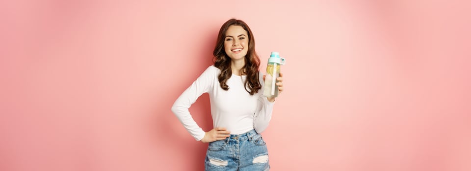 Healthy lifestyle. Happy smiling woman holding water bottle with lemon, vitamin c drink, looking joyful at camera, posing against pink background photo