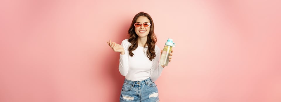 Stylish girl in spring outfit, wearing sunglasses, holding water bottle with lemon, healthy drink, laughing and smiling, standing over pink background photo