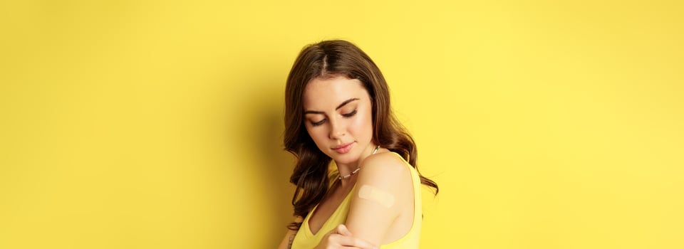 Covid-19 vaccination. Close up portrait of young millennial woman looking at patch on shoulder after vaccine from coronavirus shot, standing against yellow background photo