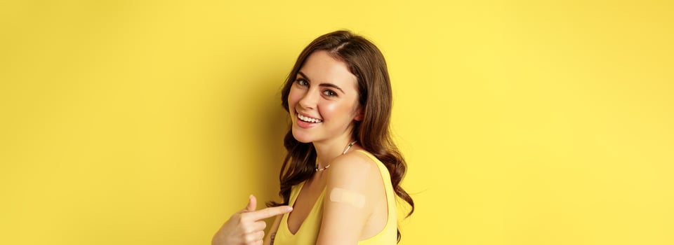 Close up of happy smiling girl with patch on shoulder after vaccination, covid-19 vaccinating campaign, standing over yellow background photo