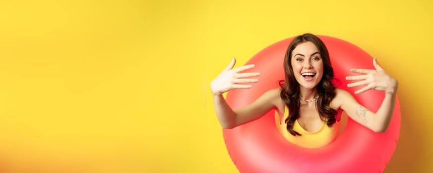 Close up portrait of beautiful happy woman inside pink swimming ring, luring, inviting people on summer vacation, beach party, standing over yellow background photo