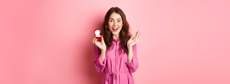 Excited young woman showing engagement ring, getting married, smiling happy, talking about wedding day, receive marriage proposal, standing over pink background photo