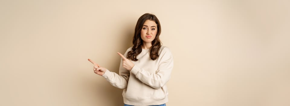 Skeptical girl looking hesitant, pointing fingers right at logo, promo text, standing over beige background photo