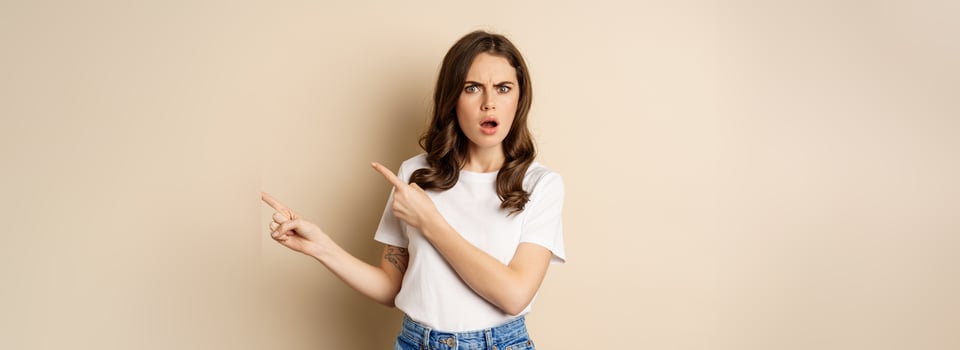 Frustrated woman looking insulted, pointing fingers right and gasping shocked, standing in t-shirt over beige background photo