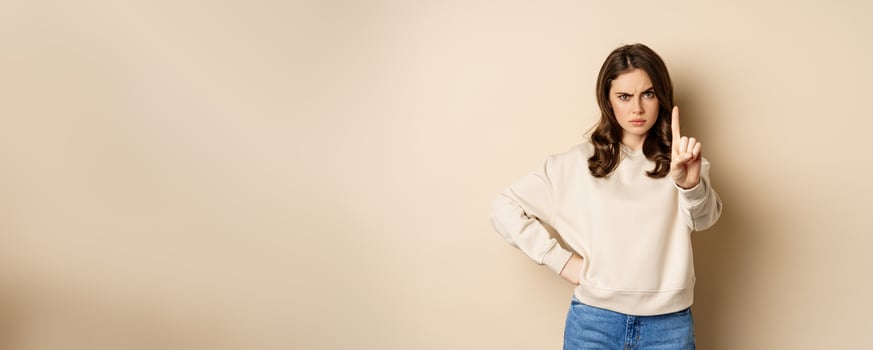 Serious woman show one finger, stop disapproval sign, forbid smth, say no, prohibit, standing over beige background photo
