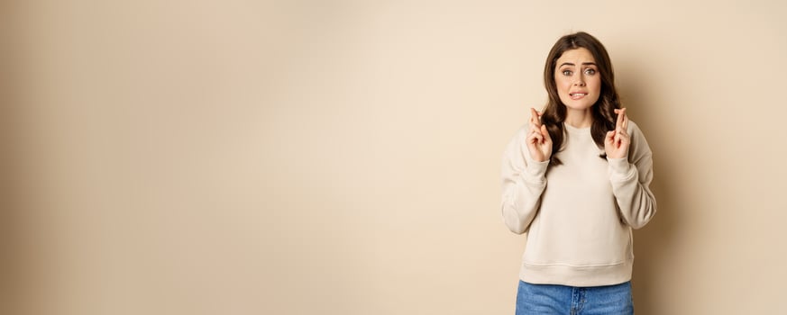 Hopeful worried girl cross fingers, making wish, standing over beige background photo