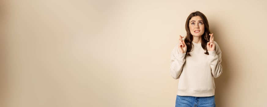 Hopeful worried girl cross fingers, making wish, standing over beige background photo