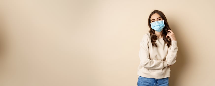 Covid-19, pandemic and quarantine concept. Beautiful modern woman in medical face mask, cross arms on chest, looking confident, beige background photo