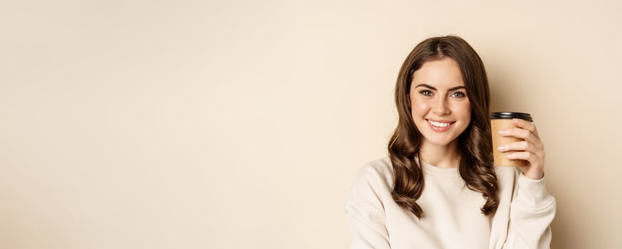 Takeaway and cafe concept. Beautiful feminine woman smiling, holding cup of coffee, posing against beige background photo