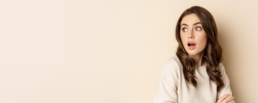 Close up portrait of brunette feminine woman turn back, look behind with surprised face expression, standing against beige background photo
