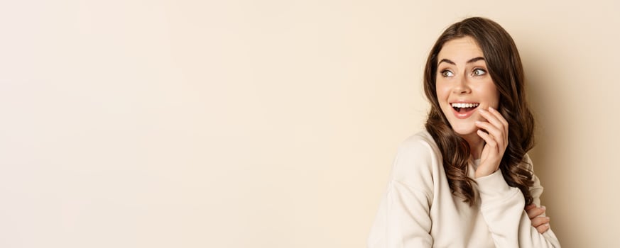 Close up portrait of brunette feminine woman turn back, look behind with surprised face expression, standing against beige background photo