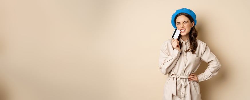 Shopping and discounts concept. Young stylish modern girl, posing with credit card, looking happy and smiling, standing over beige background photo