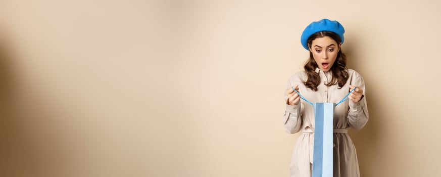 Image of girl looking surprised after opening gift bag, looking at present amazed, standing in stylish outfit over beige background photo