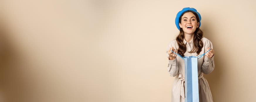 Happy young woman smiling, open shopping gift bag and laughing, giving present, standing over beige background photo