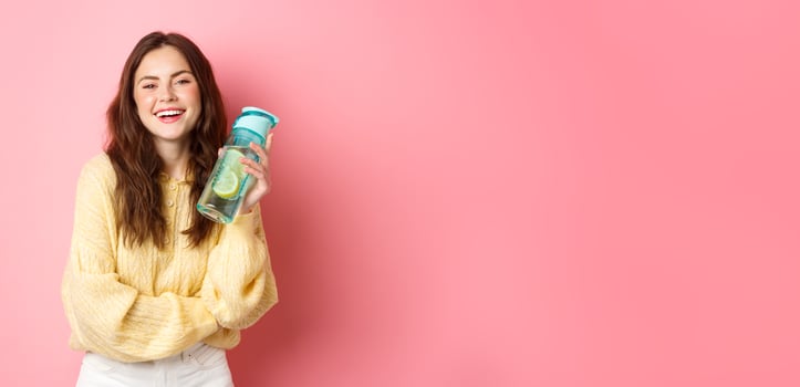 Stay hydrated. Smiling healthy girl holding water bottle with lemon and looking cheerful at camera, standing against pink studio background photo