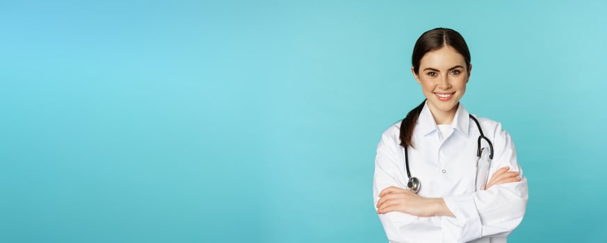 Medical staff and doctors concept. Young smiling female doctor, healthcare worker in white coat and stethoscope, looking confident, waiting patients, blue background photo