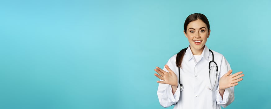 Happy and excited smiling young woman looking with excitement and surprised, standing in white lab coat and stethoscope, blue background photo