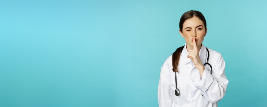 Smiling cute doctor woman, shush, press hush shh sign with finger pressed to lips, hiding secret, taboo gesture, standing over torquoise background photo