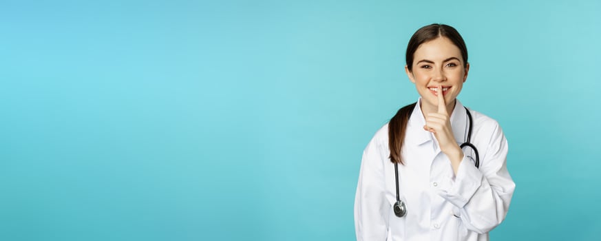 Smiling cute doctor woman, shush, press hush shh sign with finger pressed to lips, hiding secret, taboo gesture, standing over torquoise background photo