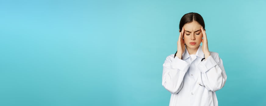 Image of woman doctor, medical personel grimacing from discomfort, having headache, migraine, feeling deezy, standing over torquoise background photo