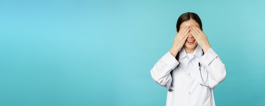 Smiling happy therapist, woman doctor cover close eyes with hands and waiting for surprised, anticipating, standing in white coat over torquoise background photo