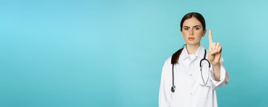 Serious looking young doctor woman, medical clinic worker showing stop, prohibition, forbidding gesture, disapprove smth, standing over torquoise background photo