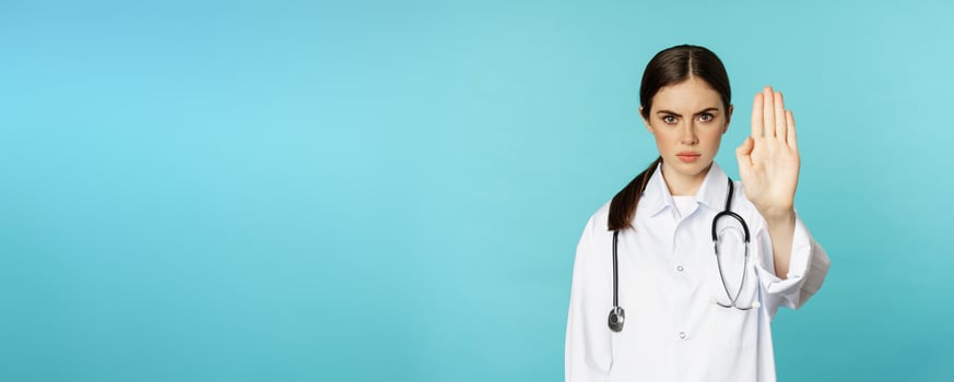 Serious looking young doctor woman, medical clinic worker showing stop, prohibition, forbidding gesture, disapprove smth, standing over torquoise background photo