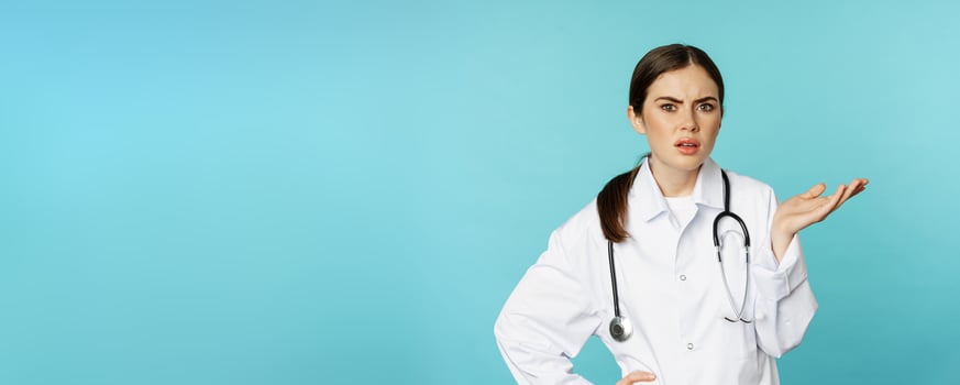 Portrait of annoyed, tired woman doctor, facepalm, roll eyes frustrated, bothered by smth stupid, standing in white coat over torquoise background photo