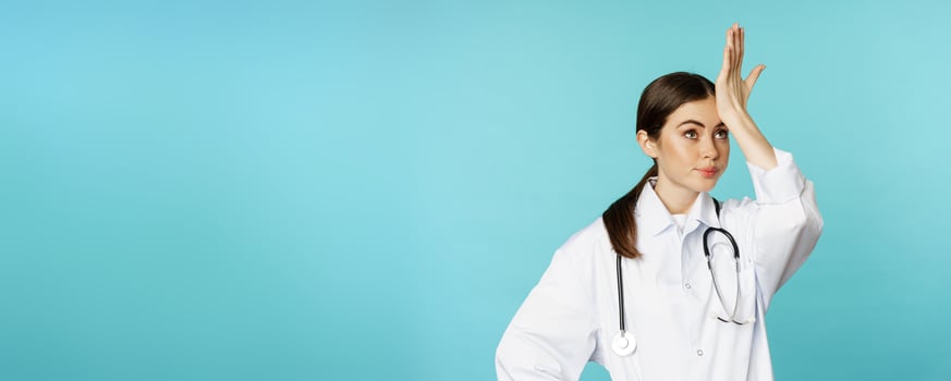 Portrait of annoyed, tired woman doctor, facepalm, roll eyes frustrated, bothered by smth stupid, standing in white coat over torquoise background photo