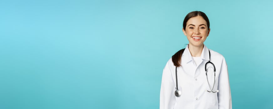 Smiling healthy medical worker, young woman doctor looking happy, standing in white coat and stethoscope against blue background photo