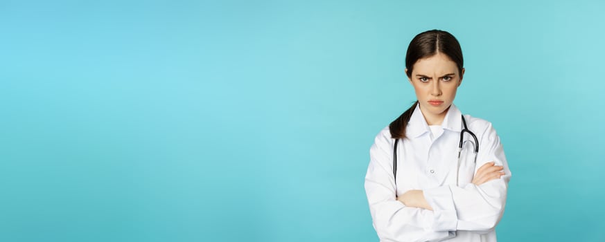 Angry doctor, female medical worker looking offended, cross arms on chest, frowning and staring annoyed at camera, standing over blue background photo