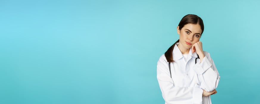 Thoughtful medical worker, young doctor woman looking unamused, bored, standing in white coat against blue background photo