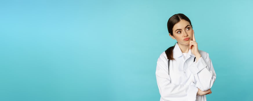 Thoughtful medical worker, young doctor woman looking unamused, bored, standing in white coat against blue background photo