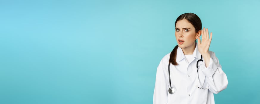 Image of confused woman doctor cant hear you, holding hand near ear and looking puzzled, speak louder gesture, torquoise background photo