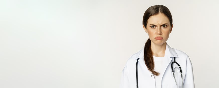 Angry and upset young woman doctor, female healthcare worker sulking, frowning disappointed, dislike smth, standing over white background photo