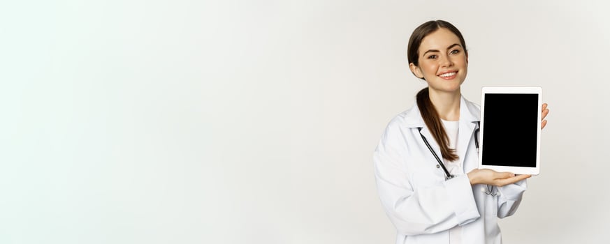 Online medical help, remote appointment. Smiling beautiful woman doctor showing digital tablet screen, demonstrating website logo, standing over white background photo